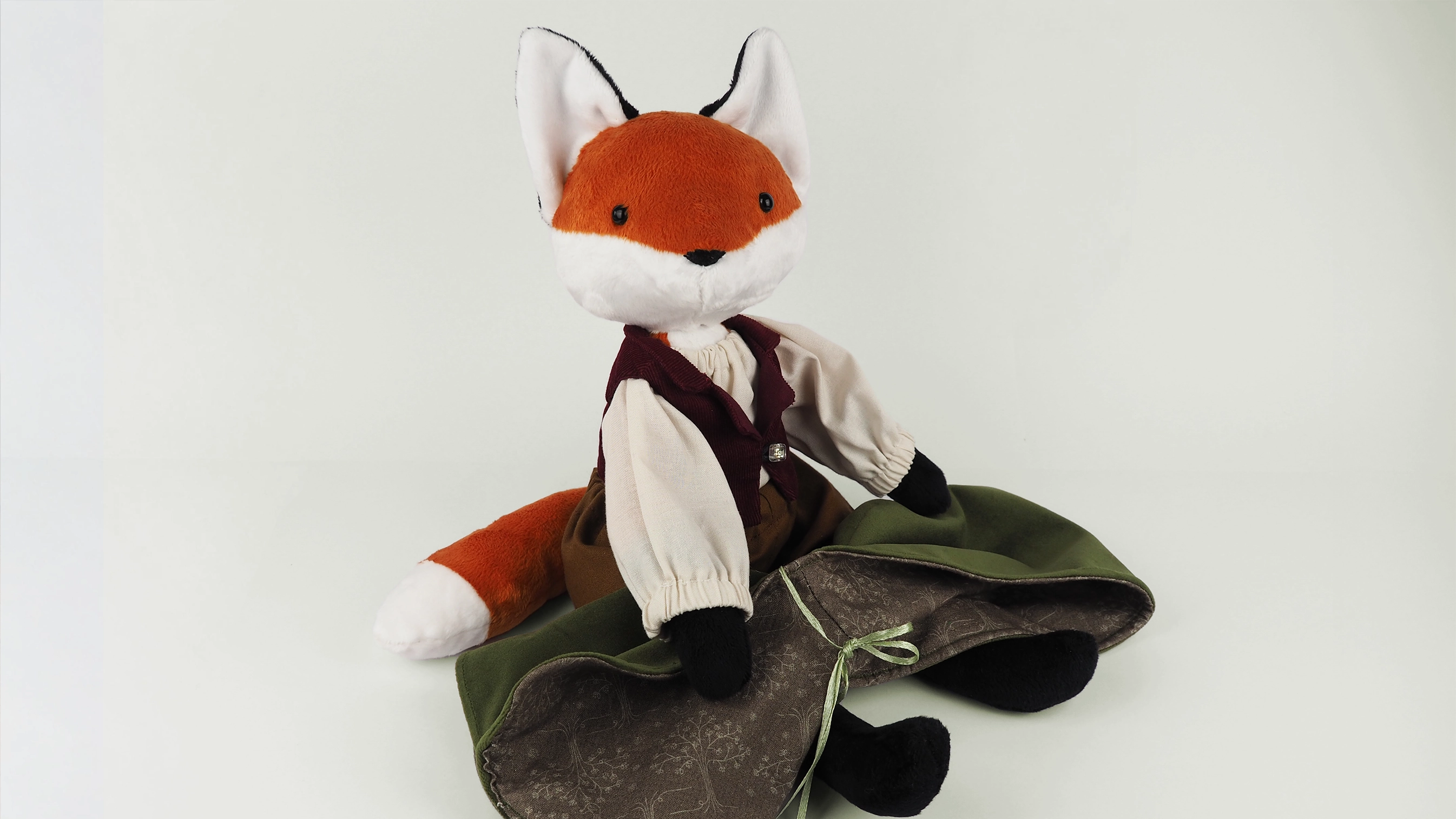 Mr Whiskers, stuffed fox doll wearing hobbit style waistcoat, shirt and trousers, with a cloak on his lap pictured against a white background.