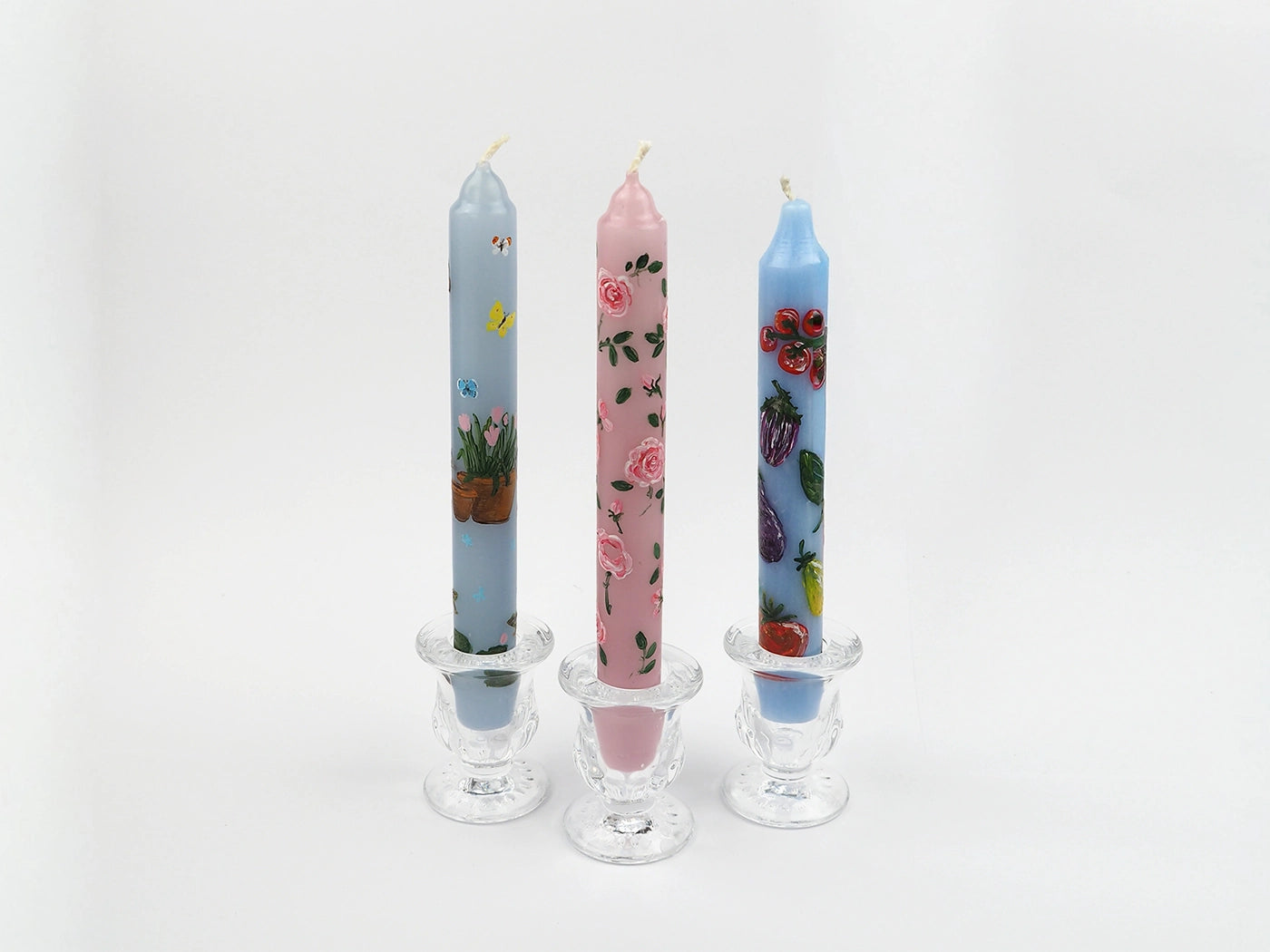 Three colorful candles with hand painted floral designs in glass candle holders on a white background